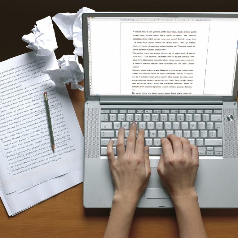 A person's hands typing on a laptop, surrounded by crumpled paper and a pencil.